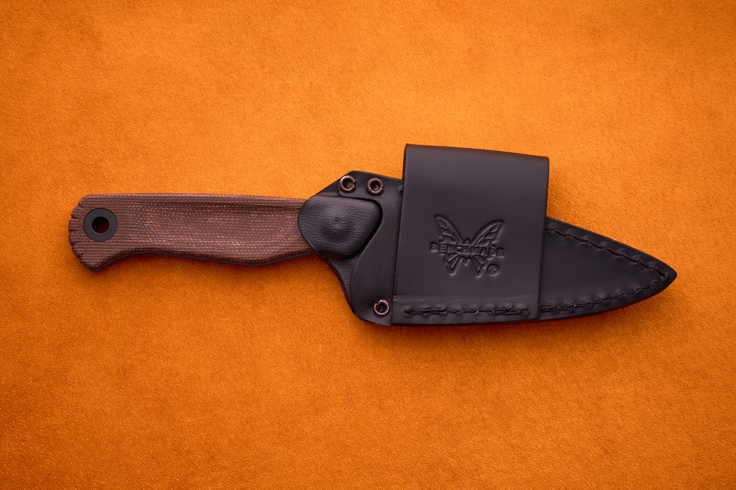 The Benchmade Dacian - Brown Micarta, a fixed-blade knife crafted from premium CPM-Magnacut steel with a textured brown micarta handle and a sleek black leather sheath adorned with the butterfly logo, rests on an orange suede background.