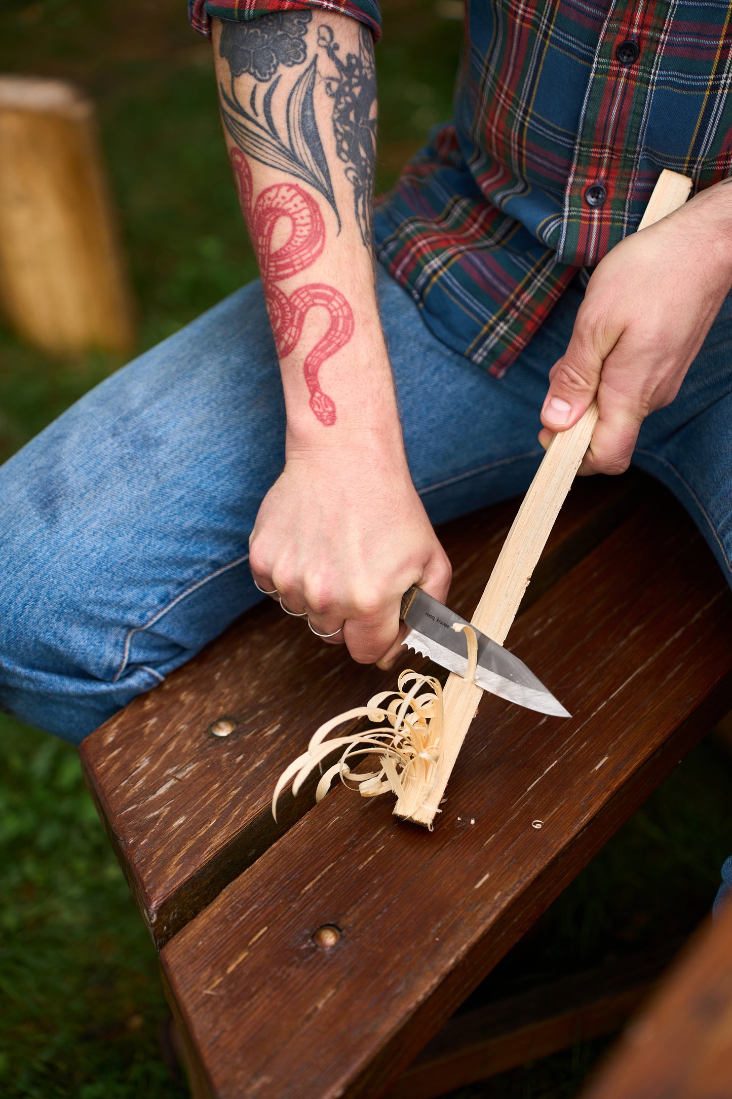 A person with a red snake tattoo on their forearm is whittling a stick with the RESQ Knives Korpi Camp Kitchen Knife while sitting on a wooden bench, wearing blue jeans and a plaid shirt.