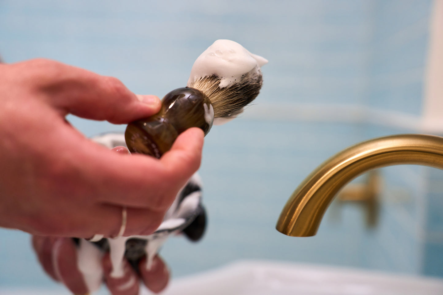 A hand holds a Kent of Inglewood Grey Badger Hair Shaving Brush covered in white shaving cream near a gold faucet, with a blue tiled wall and sink in the background—perfect for exfoliating skin and lathering hard soaps.