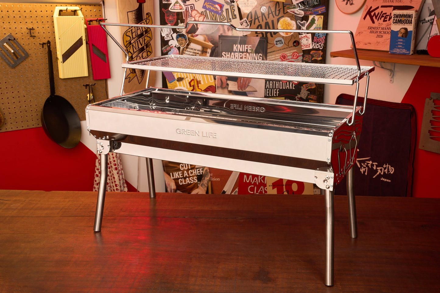 A Green Life Stainless Steel BBQ Grill stands on a wooden table in a colorful room decorated with posters, tools, and kitchen items on the walls.