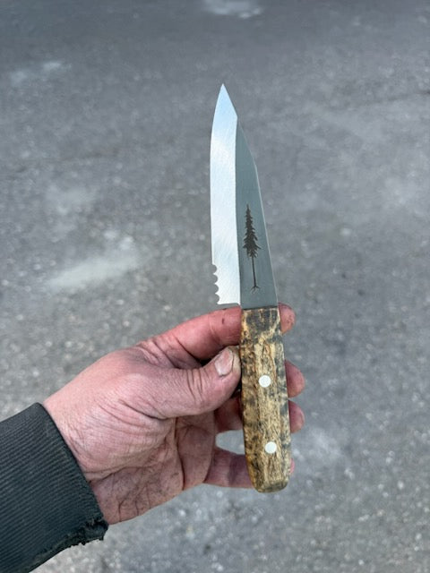 A hand holds the RESQ Knives Korpi Camp Kitchen Knife, featuring a patterned wooden handle and a shiny recycled stainless steel blade engraved with a pine tree silhouette. The background is a blurred gray surface.