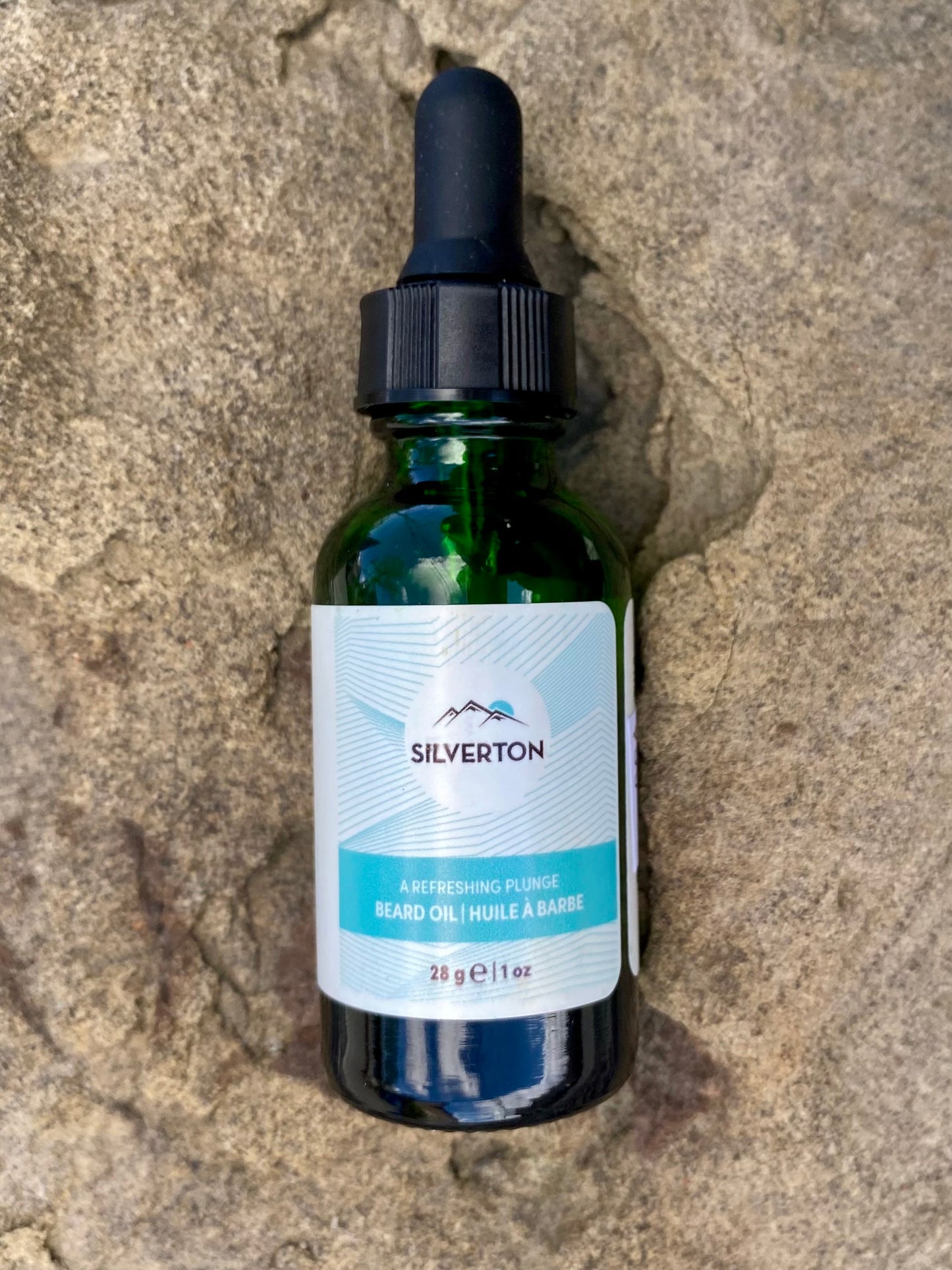 A green glass dropper bottle labeled Banff Beard Co. Silverton Falls Beard Oil rests on a textured stone surface. Its bilingual label with mountain graphics highlights premium ingredients in the 28g (1 fl oz) beard oil.