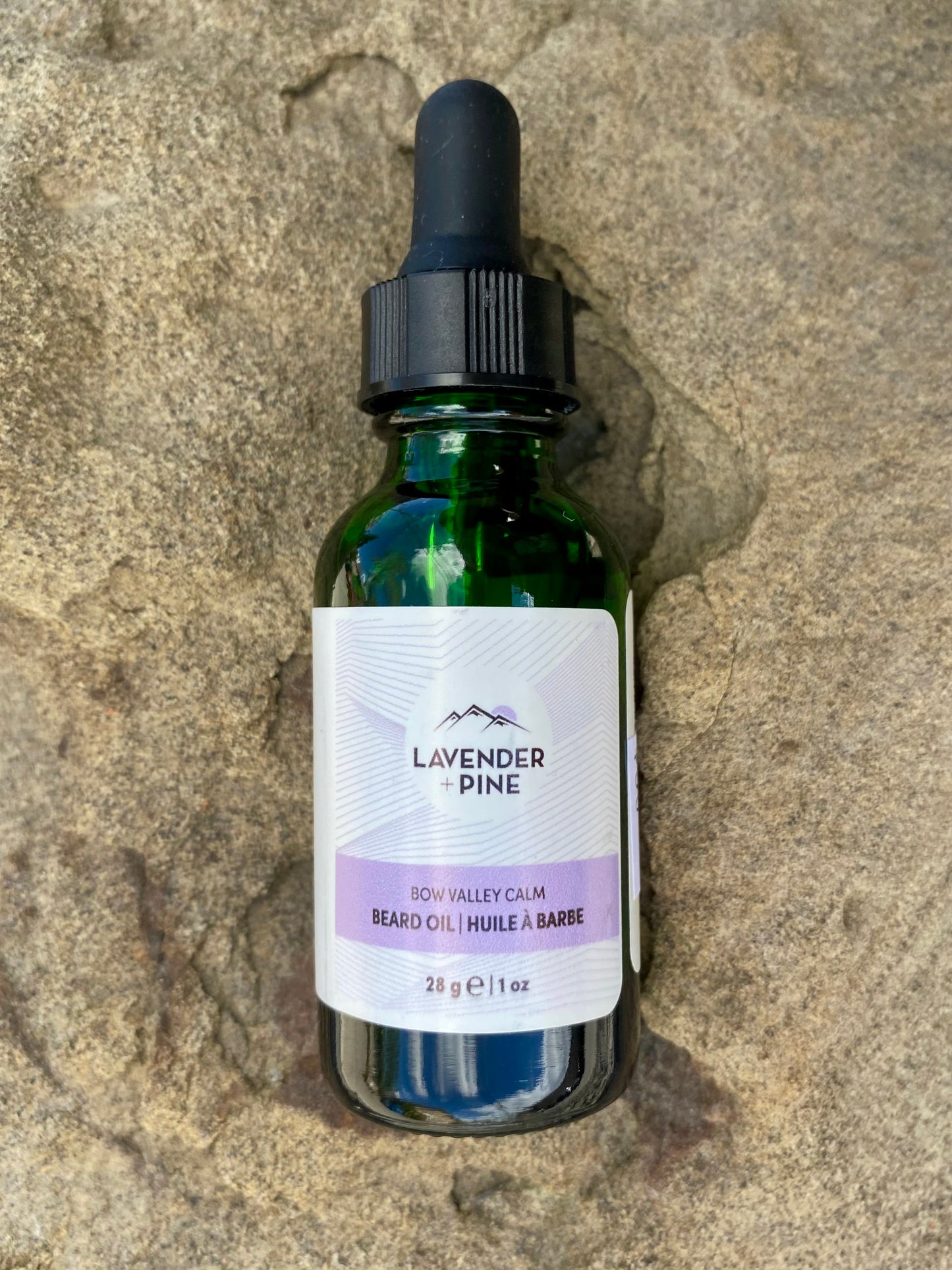 A green glass dropper bottle of Banff Beard Co. Lavender + Pine Beard Oil rests on textured stone, its white label with purple accents showing a size of 28g (1 fl oz).