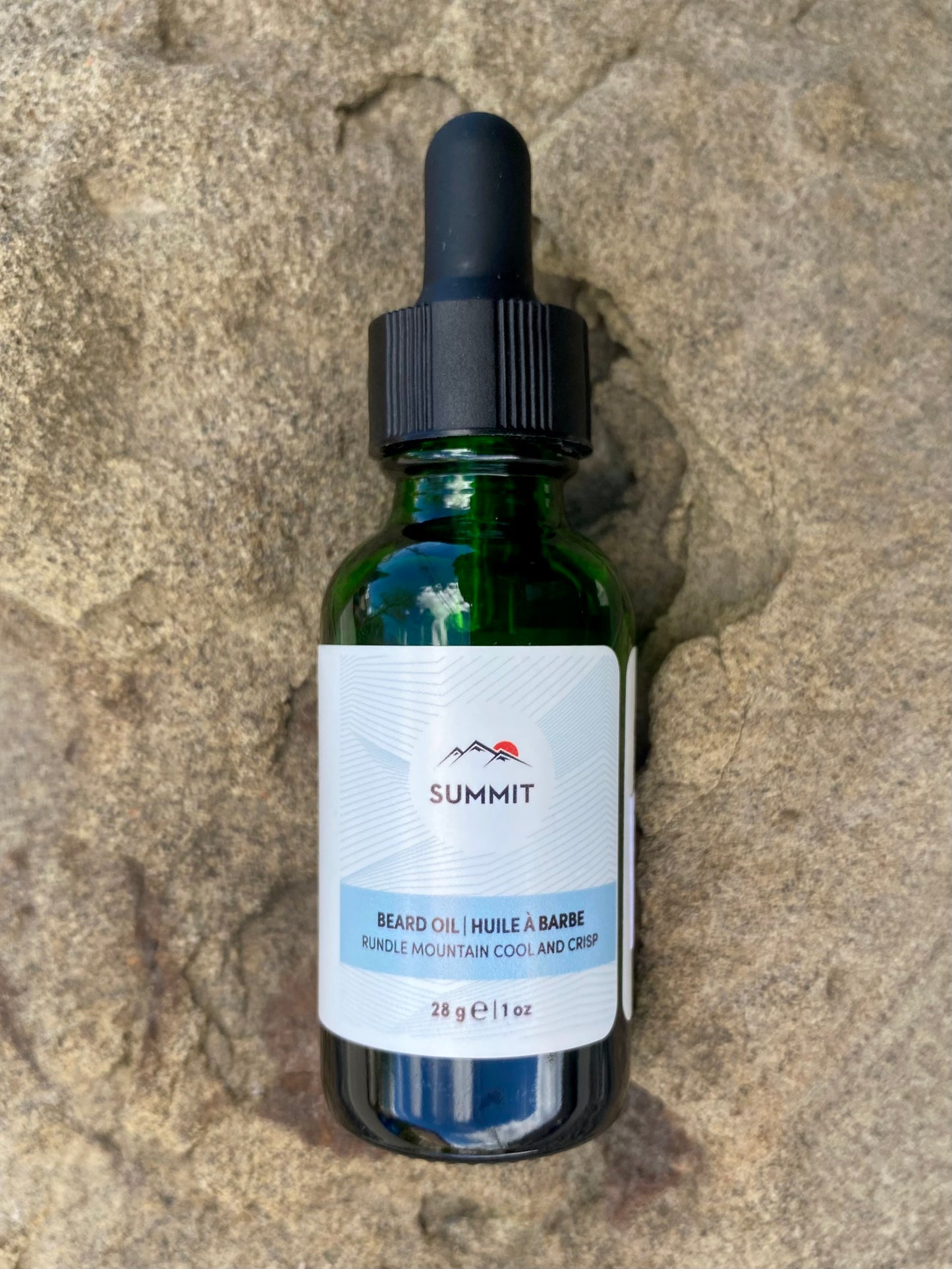 A green glass dropper bottle of Banff Beard Co. Summit Beard Oil, labeled Rundle Mountain Cool and Crisp, made with premium ingredients, sits on a textured stone surface.