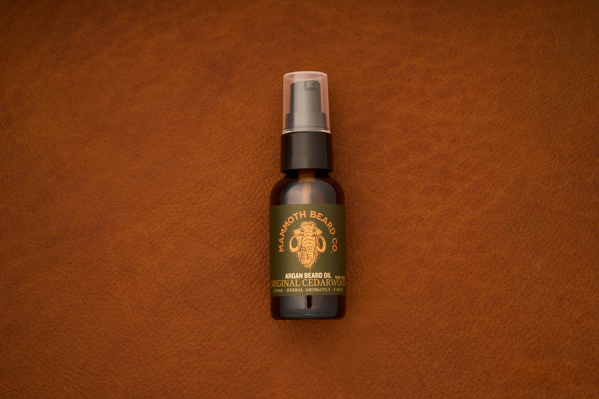 A small dark brown bottle of Mammoth Beard Co. Beard Oil - Original Cedarwood 30ml, expertly crafted with natural ingredients, sits with a spray cap on a textured brown leather surface.