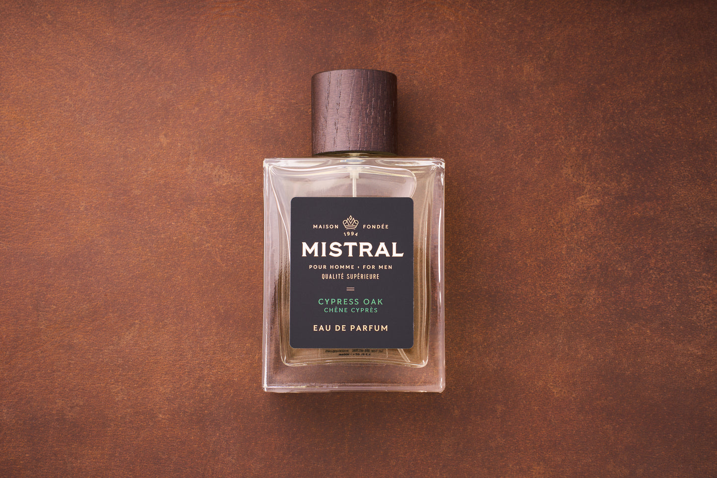 A 100ml bottle of Mistral Cypress Oak Eau de Parfum, a French men’s fragrance with a wooden cap and dark label, rests on a brown textured surface.