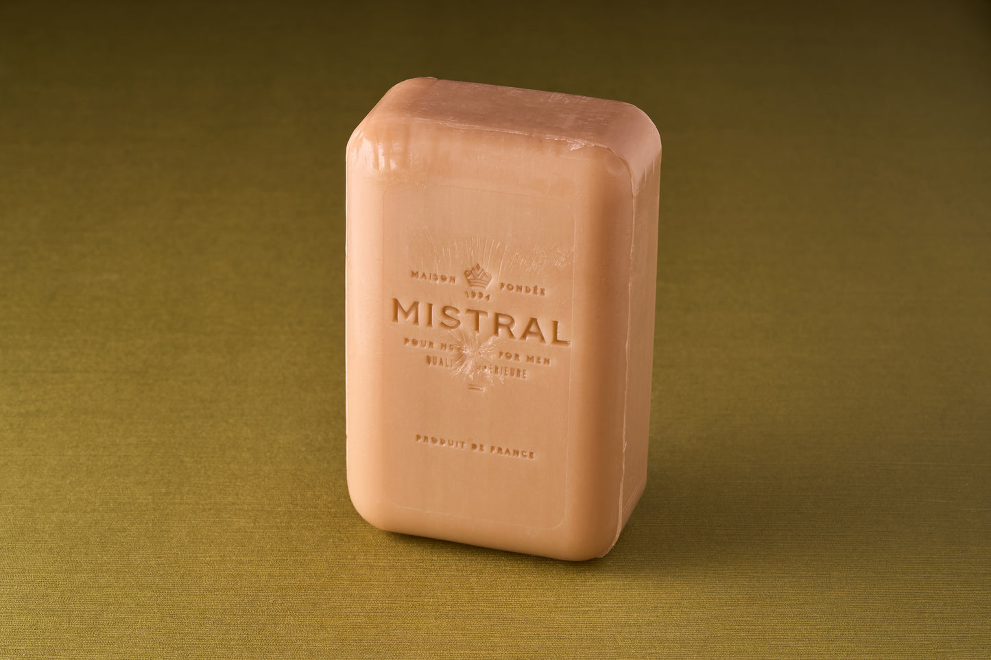 A rectangular Mistral For Men Santal Coast Bar Soap stands upright on a green surface, featuring embossed text and logo on its peach-colored front, reflecting the sophistication of Provence-inspired French fragrance.