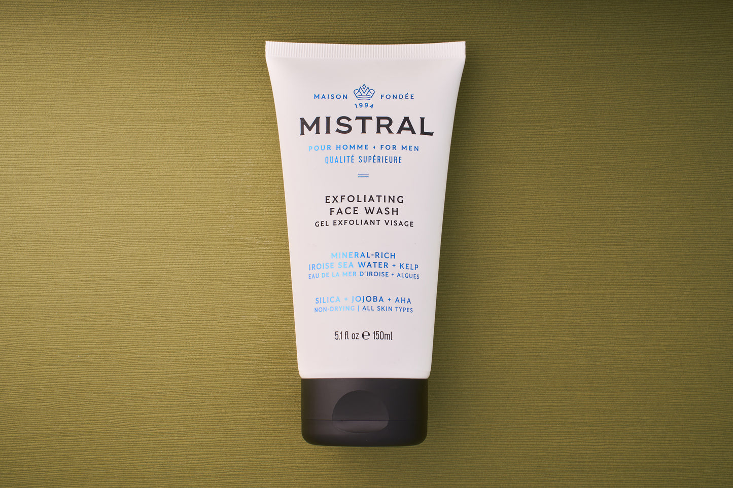 A white tube of Mistral Performance Series Exfoliating Face Wash for men, with blue and black text, sits on a textured olive-green background. Provence-inspired, it offers a French fragrance touch; details and ingredients are shown on the tube.