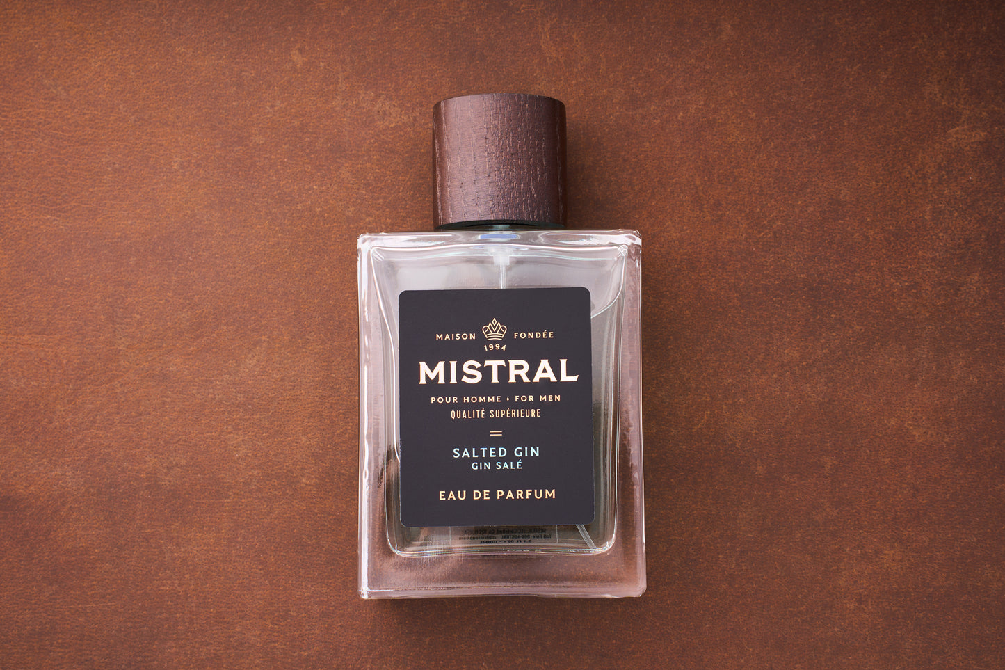 A rectangular 100ml Mistral Salted Gin Eau de Parfum bottle for men with a dark wooden cap rests on a brown surface, featuring a black label with gold and white text. This Provence-inspired fragrance is perfect for men.