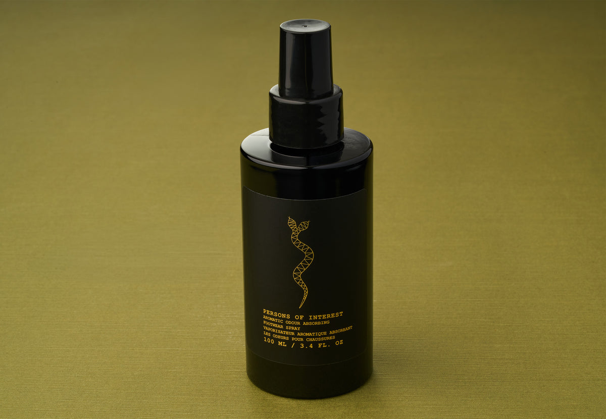 A black spray bottle labeled “Persons of Interest Aromatic Odour Absorbing Footwear Spray,” featuring gold text and a snake motif, stands on an olive green surface, hinting at an alluring fragrance inspired by rare essential oils.