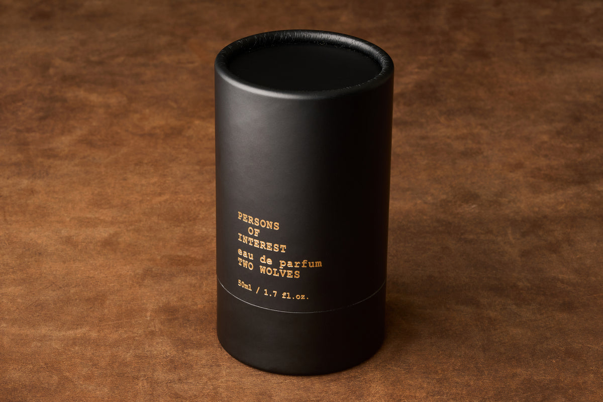A black cylindrical perfume container labeled Persons of Interest Two Wolves Eau de Parfum 50ml / 1.7 fl oz sits on a brown textured surface, evoking the allure of a light fragrance with hints of jasmine and vanilla.