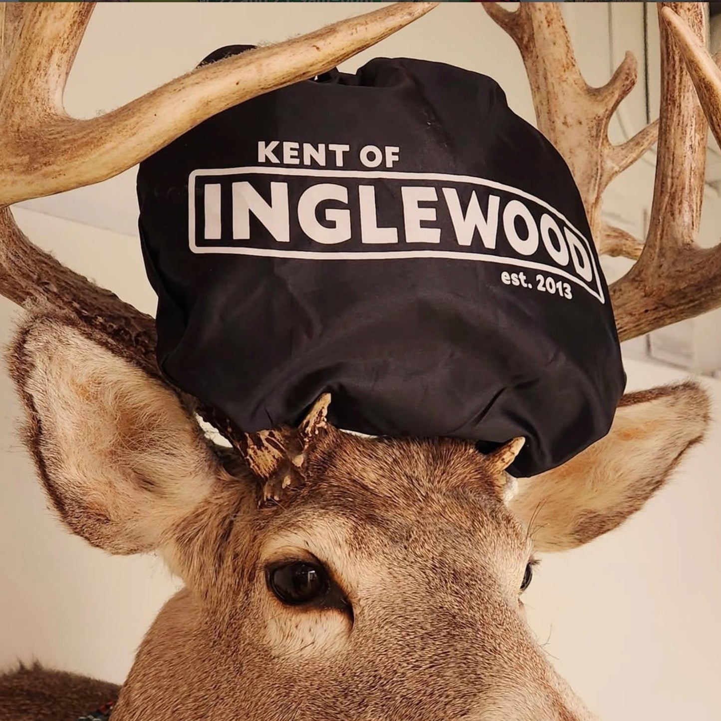 Deer head with a black bag placed on its head labeled 'Kent of Inglewood' beard bib