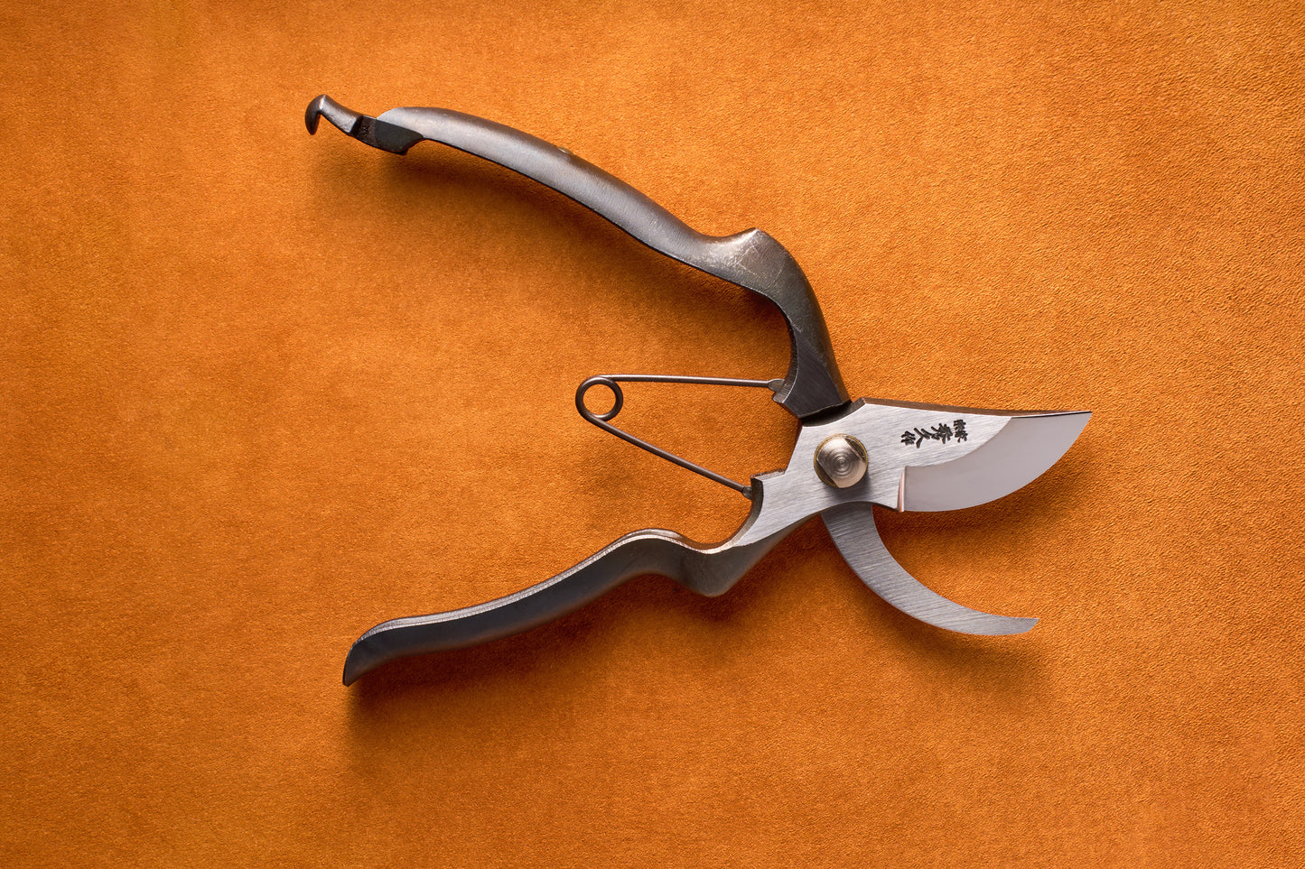 The Toyama Pruning Offset Shears 200mm T13, featuring a spring mechanism and curved blades, rest on an orange surface. Displayed open, these shears reveal sharp edges and ergonomic handles—ideal for precise work in a Japanese garden.
