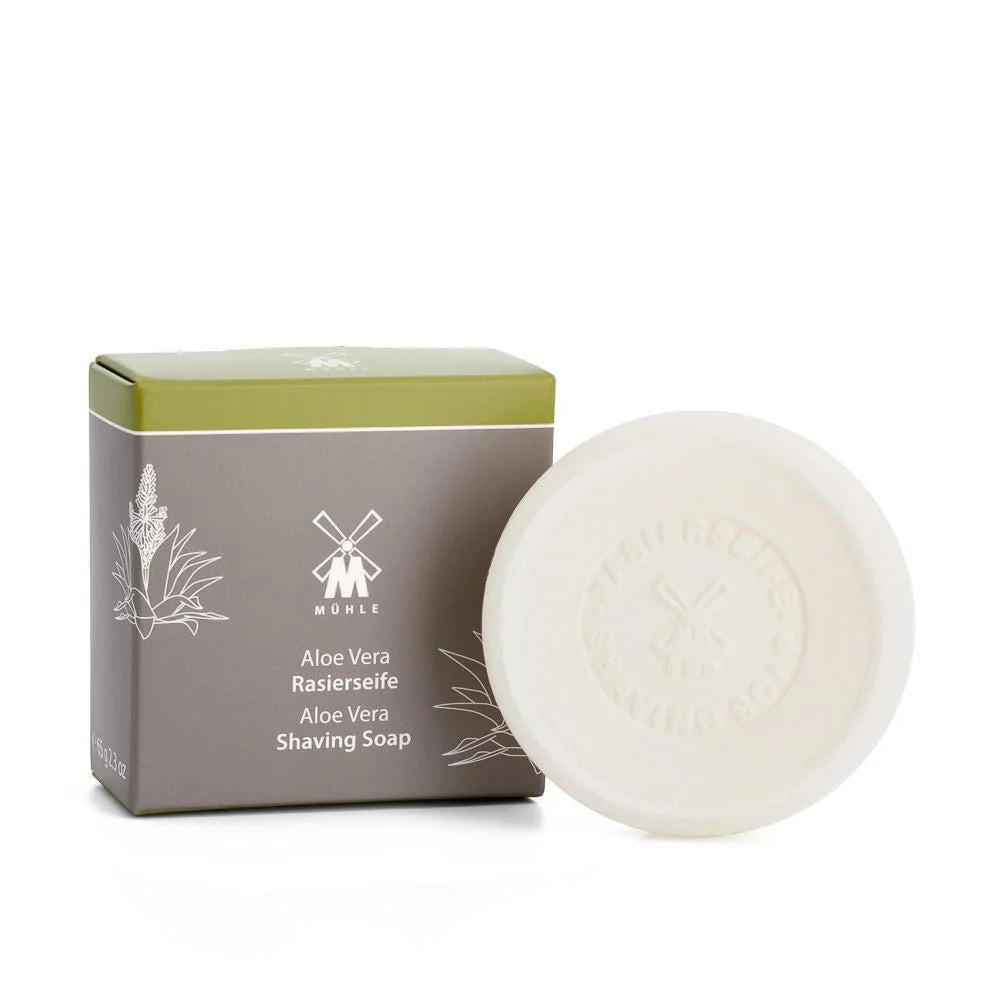 A round, white Mühle Aloe Vera Shaving Soap bar with embossed text is displayed beside a gray and green box labeled with the product name, featuring a plant illustration and the Mühle logo—perfect for sensitive skin shaving.
