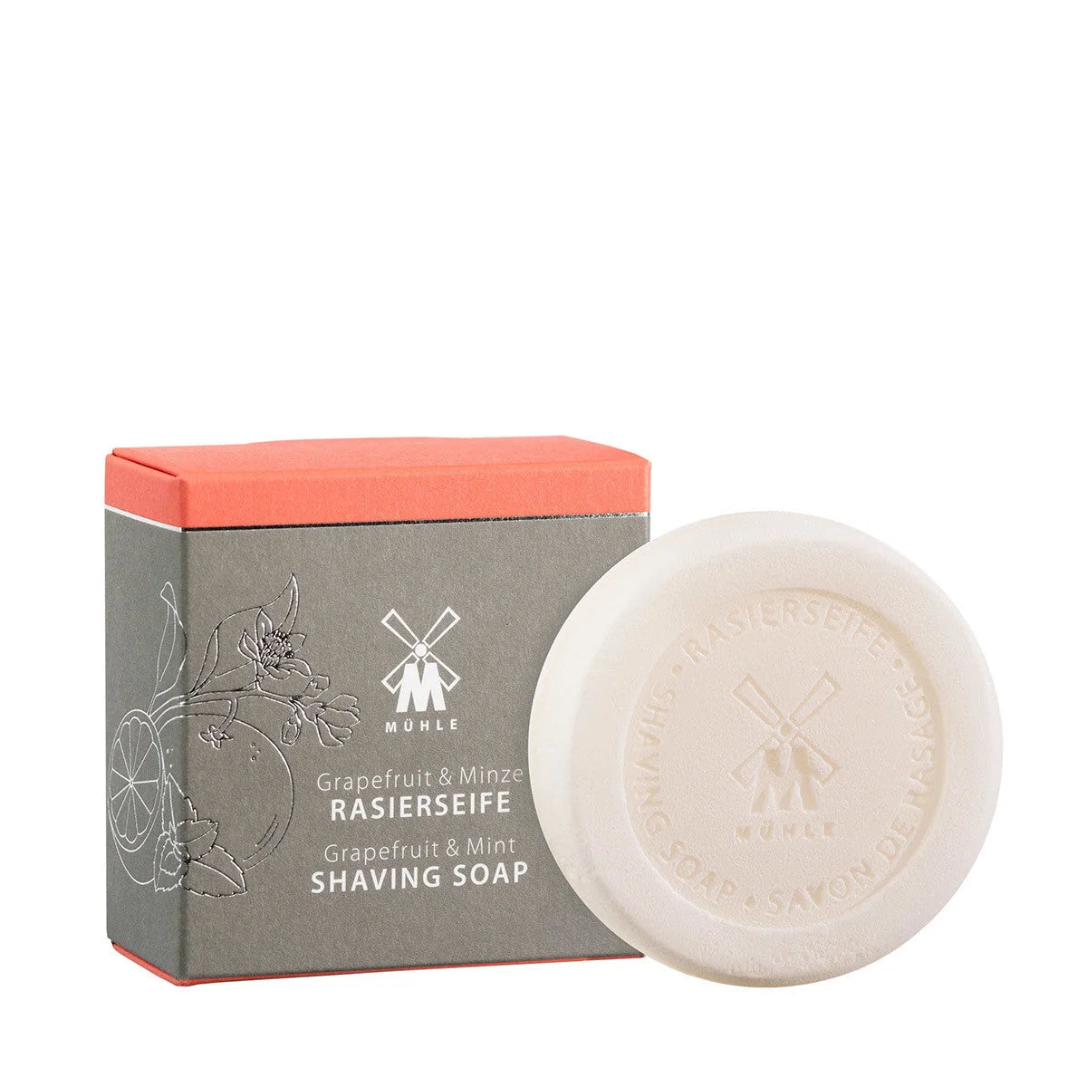 A round white shaving soap featuring embossed text sits in front of a gray and orange box decorated with floral and fruit motifs. The packaging displays “Mühle Grapefruit & Mint Shaving Soap” in both English and German, emphasizing this vegan soap’s luxurious and refreshing grapefruit mint scent.