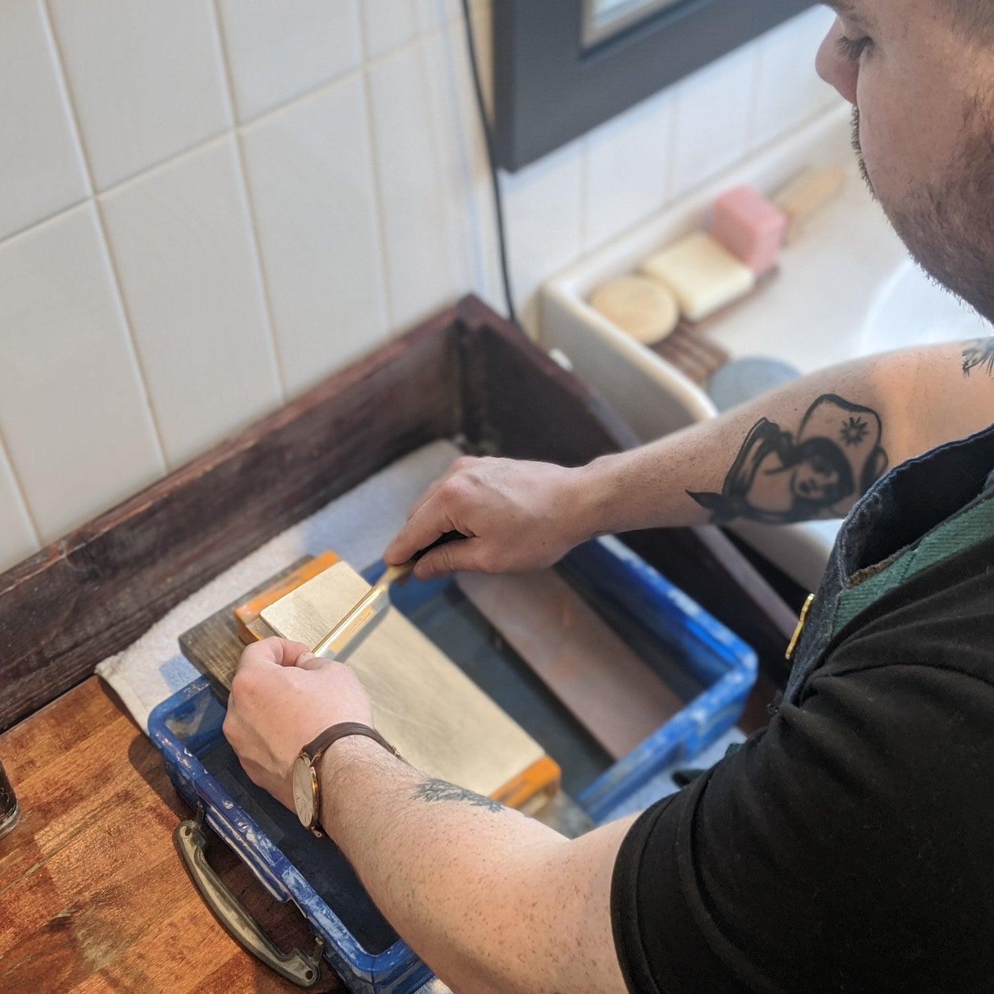 In-store Razor Honing service at Kent of Inglewood, featuring a tattooed individual honing a straight razor on a wet whetstone in a sink area.