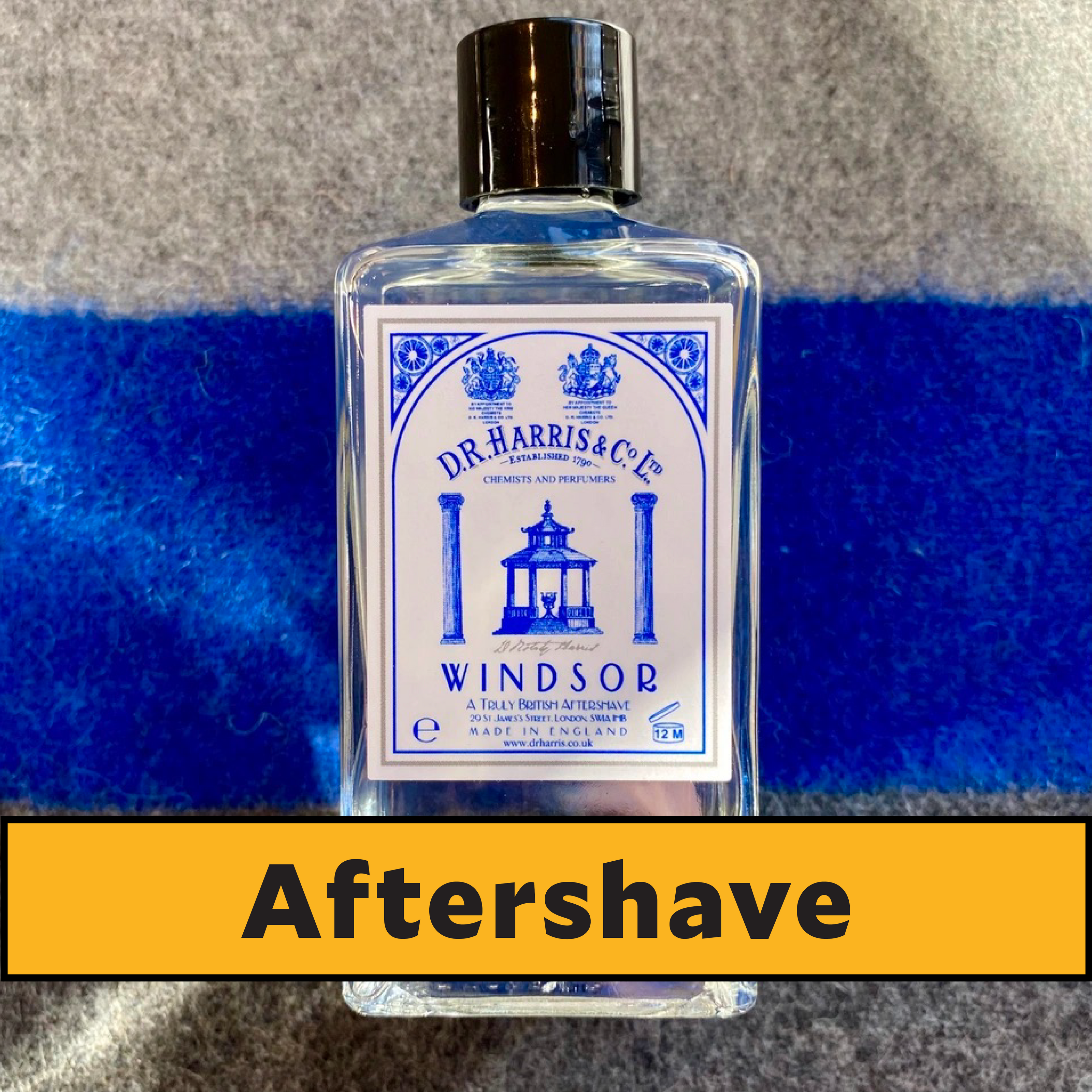A bottle of Dr. Harris & Co. Windsor aftershave sits on a blue-striped surface, with a yellow label below reading Aftershave in bold black letters.