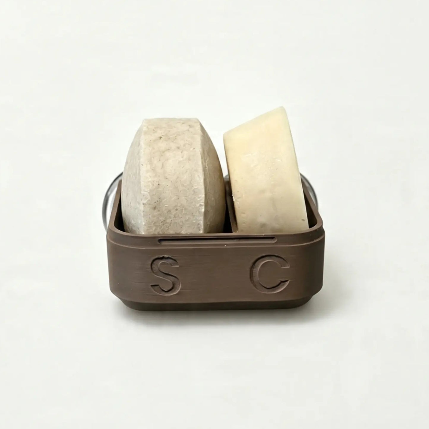 Two bars—one beige and one off-white—are displayed in The High End Hippie 2-in-1 Shampoo and Conditioner Case, a brown container with the letters S and C embossed on the front, set against a plain white background. This stylish case is perfect for sustainable shower storage, keeping your essentials organized while adding an elegant touch to your bathroom.
