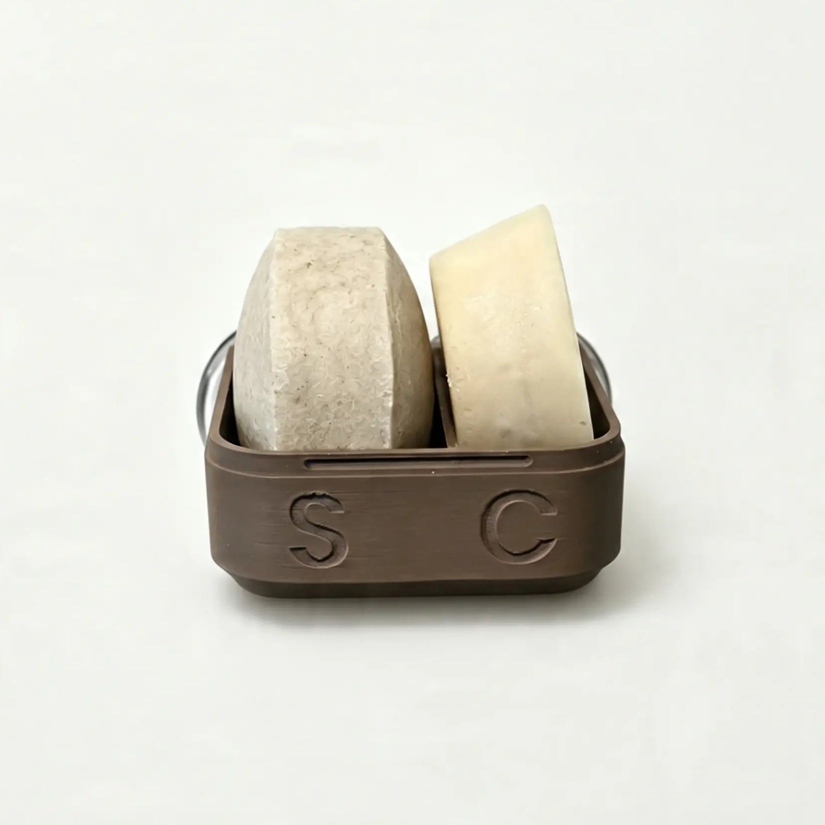 Two bars—one beige and one off-white—are displayed in The High End Hippie 2-in-1 Shampoo and Conditioner Case, a brown container with the letters S and C embossed on the front, set against a plain white background. This stylish case is perfect for sustainable shower storage, keeping your essentials organized while adding an elegant touch to your bathroom.