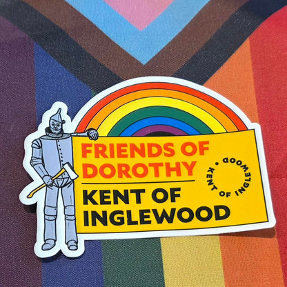 The Kent of Inglewood Pride Sticker features the Tin Man holding a rainbow sign that reads "FRIENDS OF DOROTHY, KENT OF INGLEWOOD" against a vibrant multicolored Pride flag background. This colorful sticker celebrates both Kent of Inglewood and Skipping Stone Calgary, making it an ideal addition to any collection in support of the LGBTQ+ community.