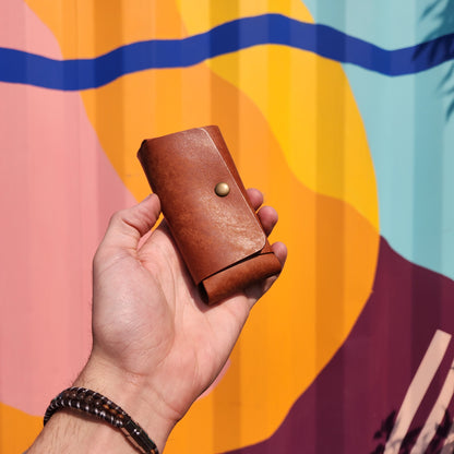 Hand holds Jack Raymond Leather Safety Razor Holster, a small brown-leather pouch with brass snap, against an abstract mural, reflecting classic shaving elegance from Kent of Inglewood.