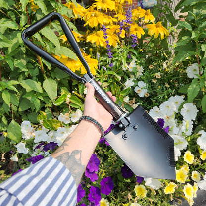 Hand holding a Captain Stag Foldable Steel Shovel against a floral backdrop, showcasing its compact design, perfect for camping or emergency use.