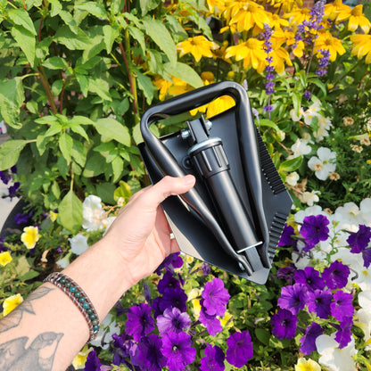 Hand holding Captain Stag Foldable Steel Shovel over blossoming flower bed, showcasing its compact, hinged design ideal for camping or roadside emergencies.