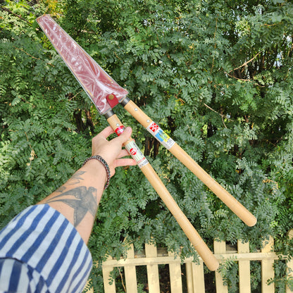 Tattooed forearm reaching for Yoshioka Shirogami Hedge Shears, showcasing wooden handles and covered blades, against a leafy hedge and picket fence backdrop.