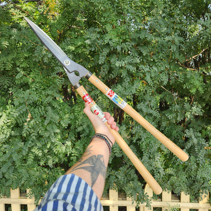 Yoshioka Shirogami Hedge Shears with 180mm blades and 450mm wooden handles, held by a tattooed arm in front of a hedge.