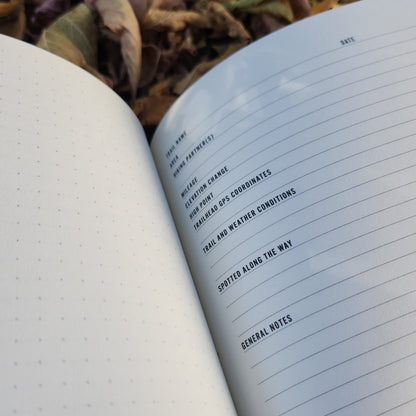 Trail Notes hiking-log notebook open on dried leaves, featuring a dotted grid page for sketches and a detailed log page for documenting trail adventures.