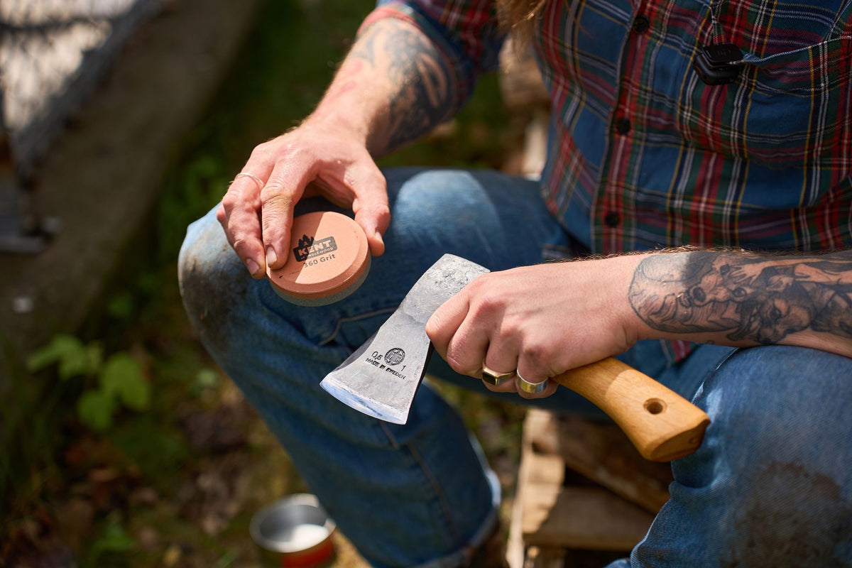A person with tattoos uses the Camp Kent of Inglewood Axe Sharpening Stone to sharpen a hatchet while sitting outdoors on a wooden surface. They are wearing a plaid shirt and jeans, with greenery visible in the background.