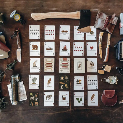 Bradley Mountain Survival Playing Cards spread on a table with outdoor tools like a hatchet, compass, and knives, showcasing wilderness-themed illustrations and survival tips.