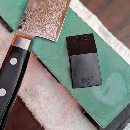 Knifewear Sharpening Angle Guide with 15° and 20° markers beside a Damascus-style chef's knife on a green whetstone, illustrating precise sharpening angles.