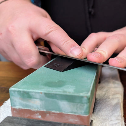 Close-up of hands sharpening a knife with Knifewear Sharpening Angle Guide on a whetstone, ensuring a precise 20° angle for optimal blade maintenance.