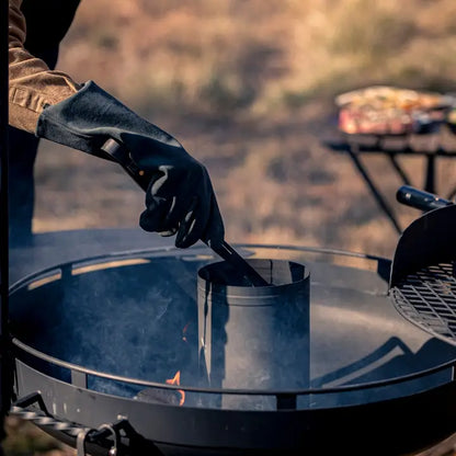 A person uses Barebones Open Fire Gloves to safely handle coals in a kettle grill, showcasing their heat resistance and ease of use.
