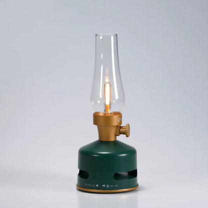 The KooKoo MoriMori Light & Sound Lantern is a modern LED lantern with a nostalgic design, featuring a green and gold base, clear glass chimney, and visible faux flame, set against a plain light gray background.