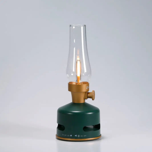 The KooKoo MoriMori Light & Sound Lantern is a modern LED lantern with a nostalgic design, featuring a green and gold base, clear glass chimney, and visible faux flame, set against a plain light gray background.