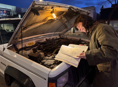 Person using Barebones Edison Light Stick under SUV hood, reading a repair manual, highlighting its dual function as an outdoor lantern and emergency flashlight.