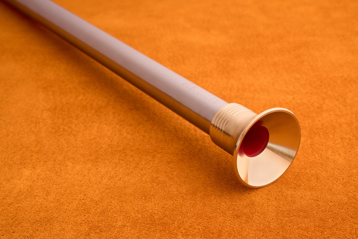 The Barebones Telescoping Fire Blower, a metallic trumpet-like tool featuring a gold bell and red interior, rests on an orange textured surface.