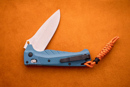 A Benchmade Adira folding pocket knife with a blue handle and a partially open silver MagnaCut blade, featuring an orange paracord lanyard, is placed on an orange textured surface.