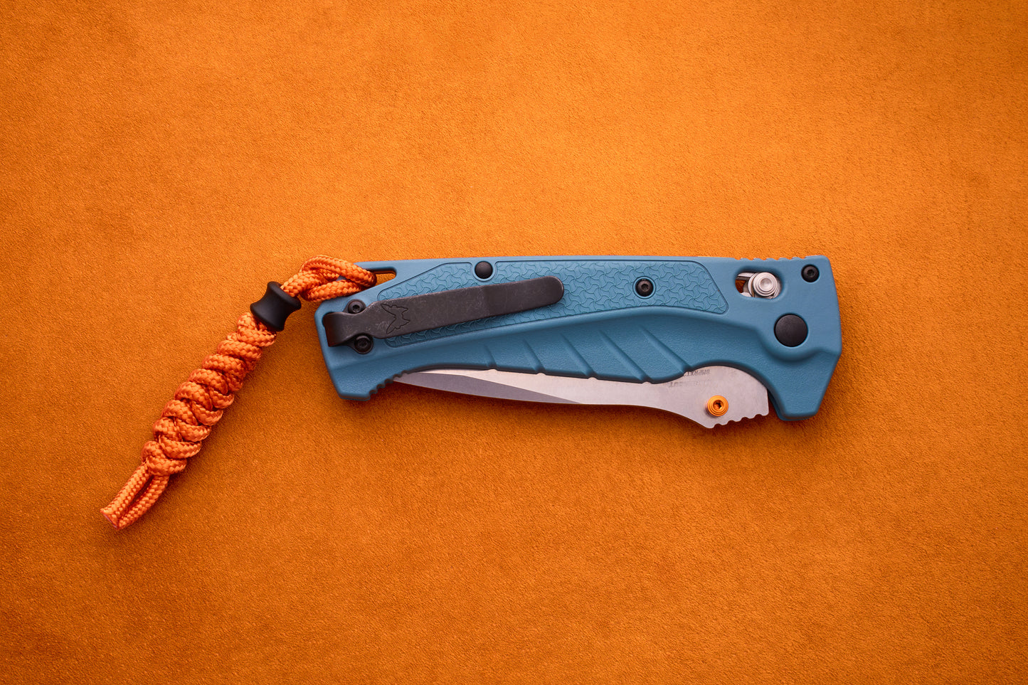 A blue Benchmade Adira folding pocket knife with a partially visible MagnaCut blade and an orange paracord lanyard rests on an orange suede surface.