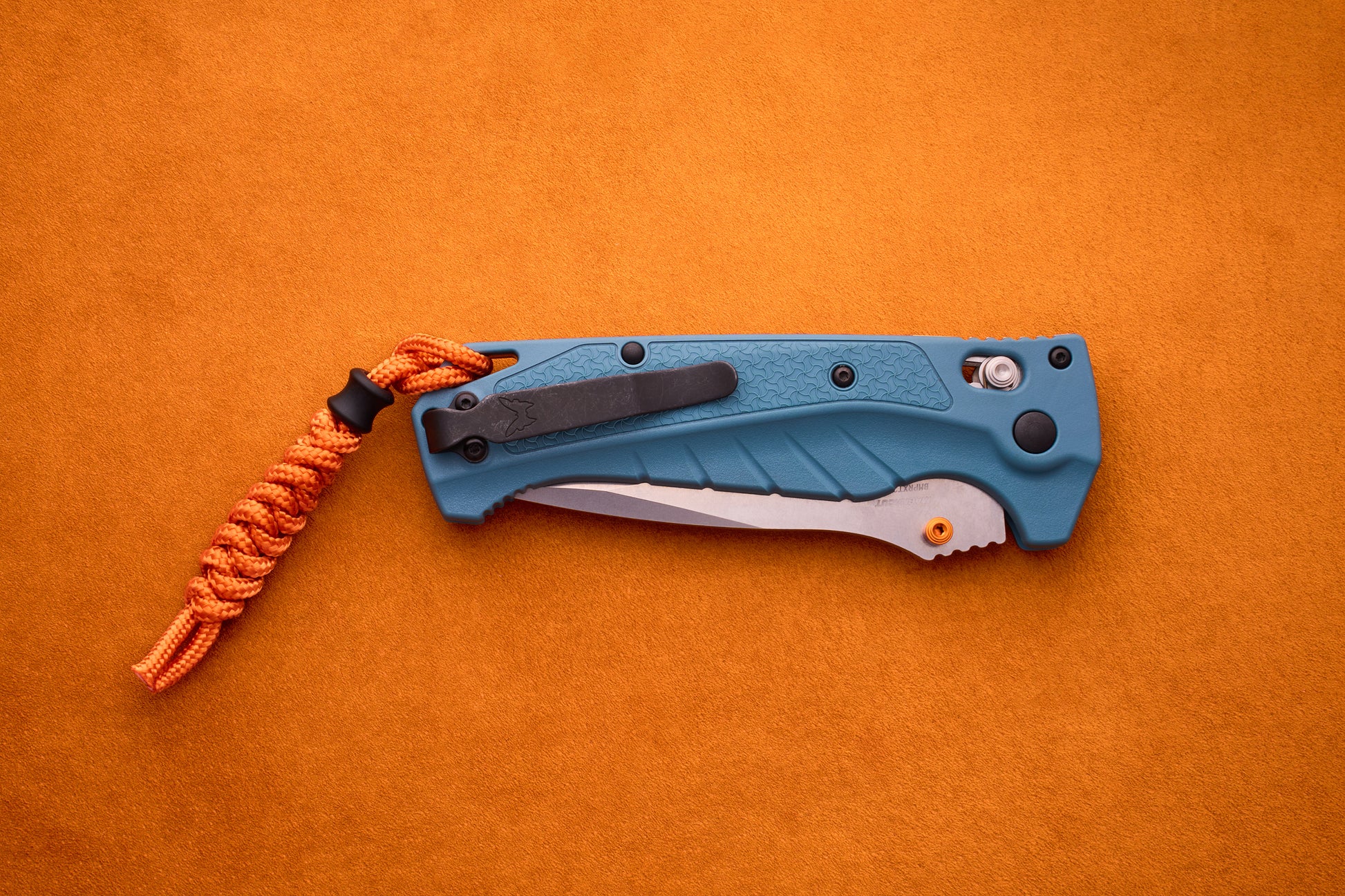 A blue Benchmade Adira folding pocket knife with a partially visible MagnaCut blade and an orange paracord lanyard rests on an orange suede surface.