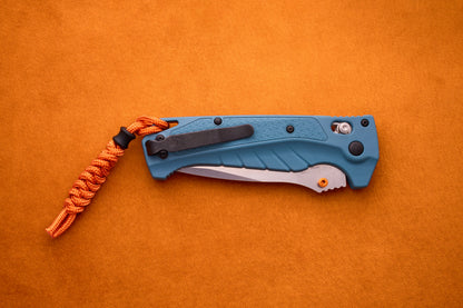 A blue Benchmade Adira folding pocket knife with a partially visible MagnaCut blade and an orange paracord lanyard rests on an orange suede surface.