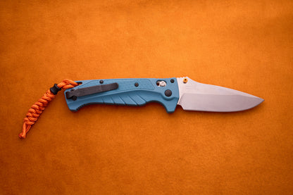 A Benchmade Adira folding knife with a blue handle and silver MagnaCut blade, featuring an orange lanyard, is placed on an orange textured background.