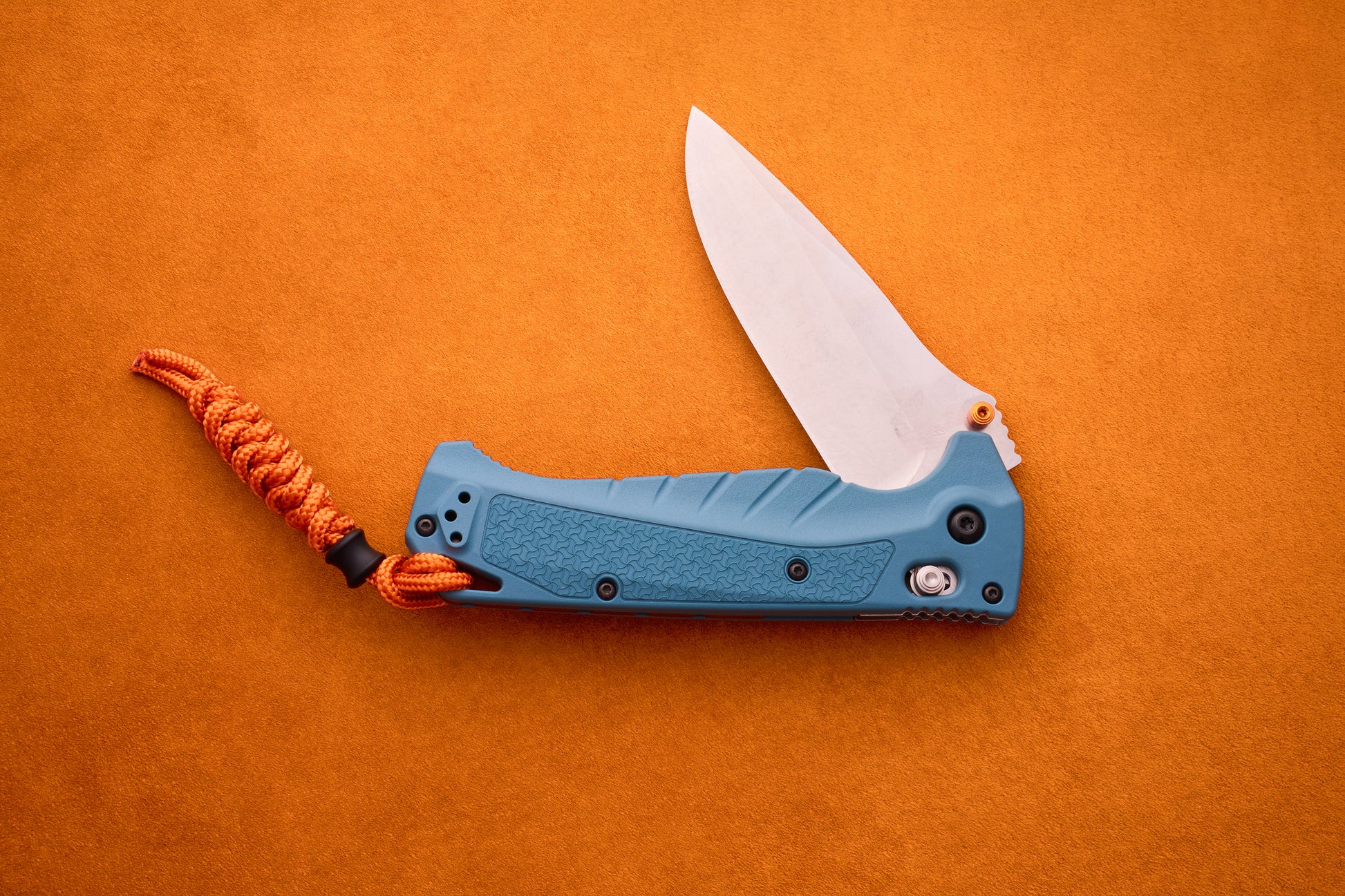 A blue Benchmade Adira folding pocket knife with an open MagnaCut blade and an orange paracord lanyard rests on an orange textured surface.