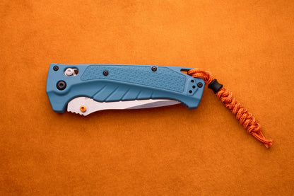 The Benchmade Adira, a blue folding pocket knife with a textured handle and an orange paracord lanyard, featuring a high-performance MagnaCut blade, is placed on an orange surface.