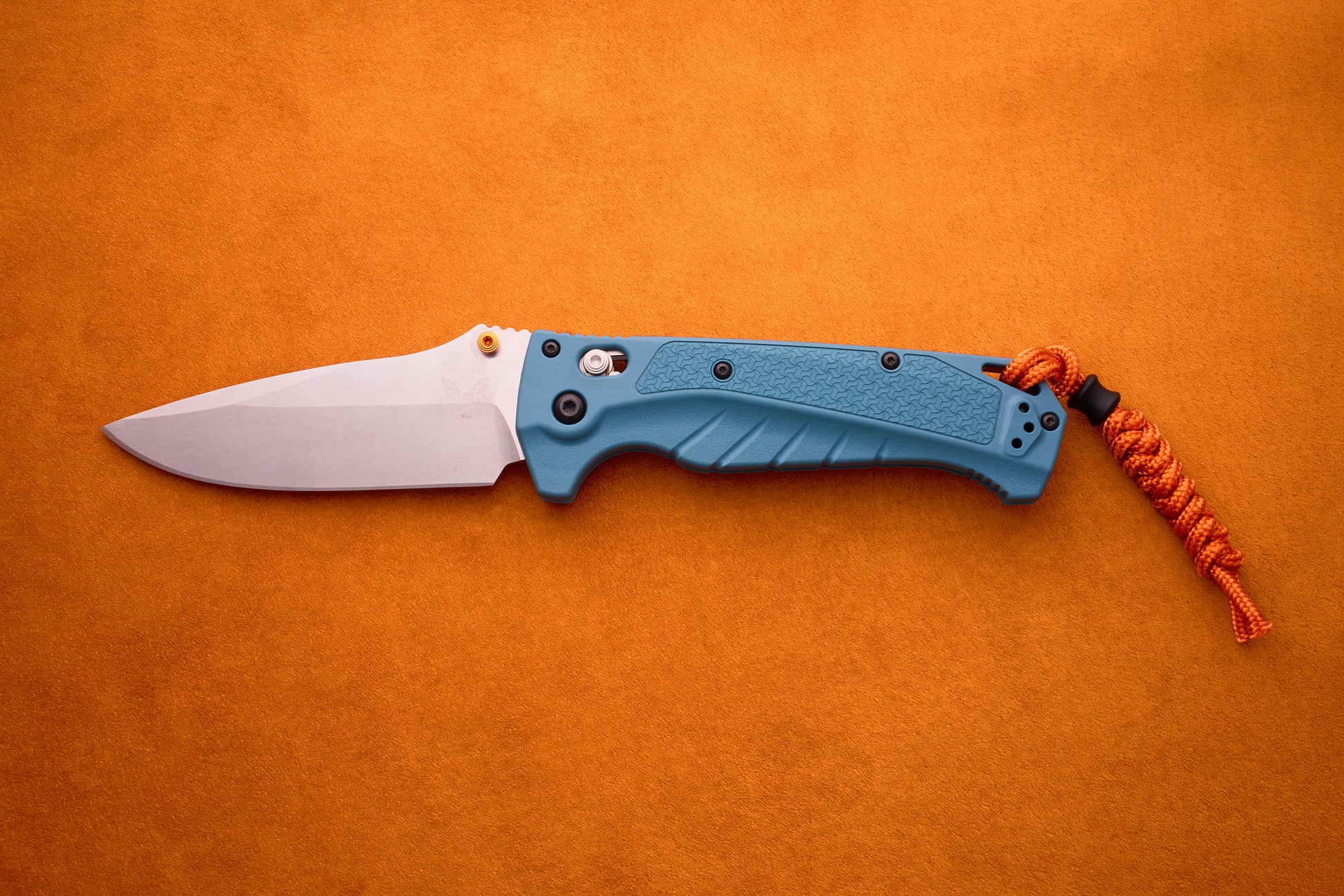 A Benchmade Adira folding knife with a MagnaCut silver blade, blue handle, and orange paracord lanyard is displayed on an orange surface.