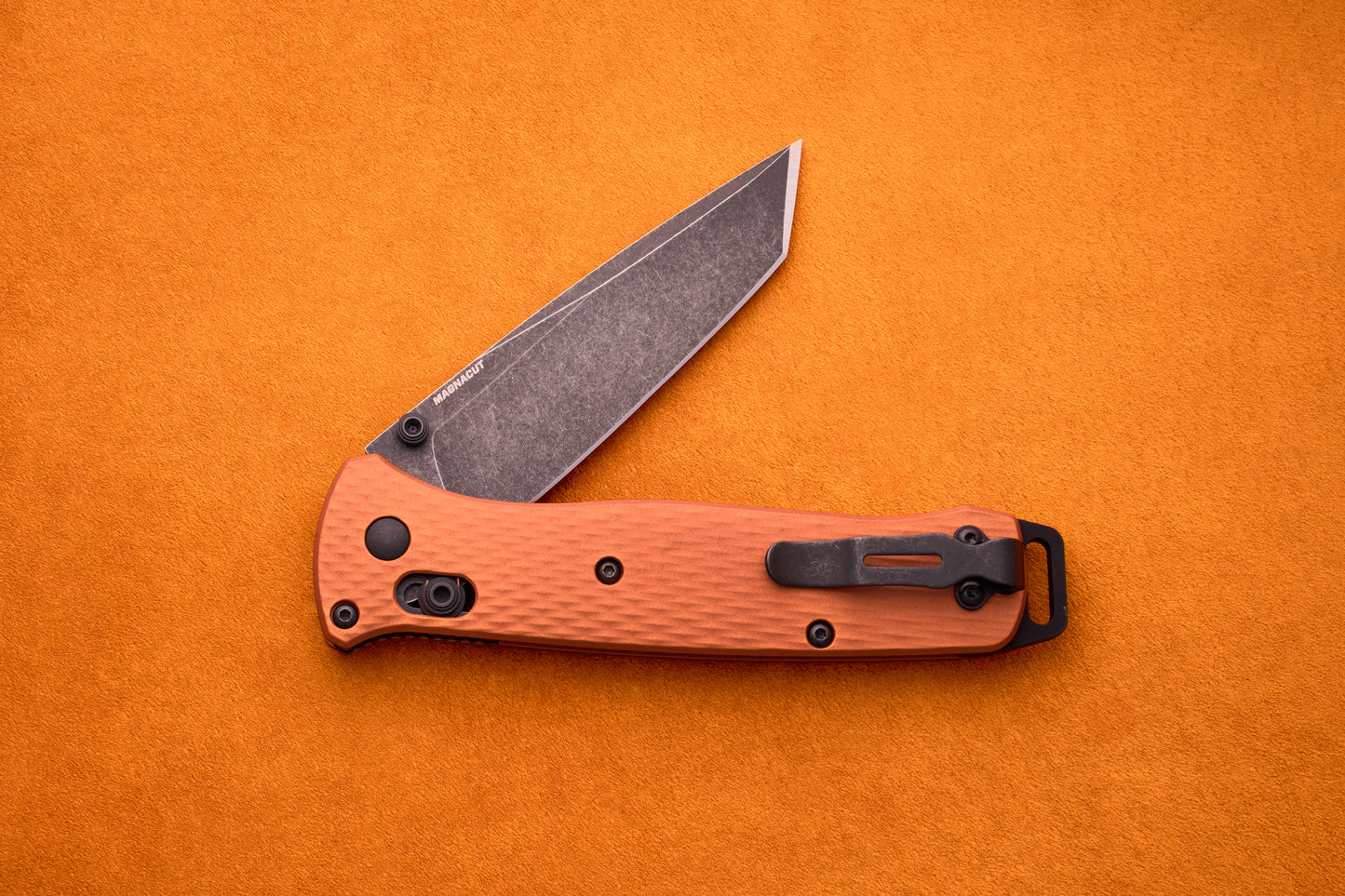 The Benchmade Bailout, featuring a textured orange handle and a partially open, gray CPM-Magnacut blade, is displayed on an orange background.