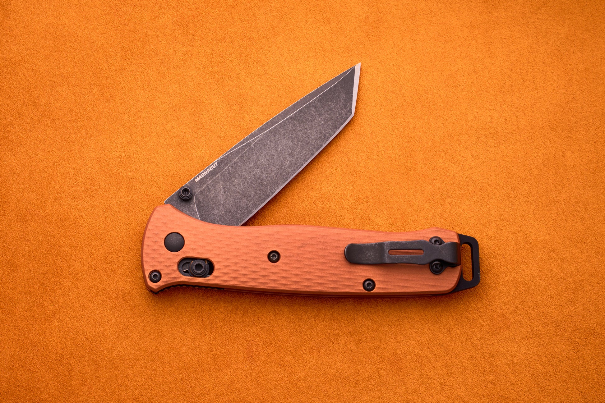 The Benchmade Bailout, featuring a textured orange handle and a partially open, gray CPM-Magnacut blade, is displayed on an orange background.