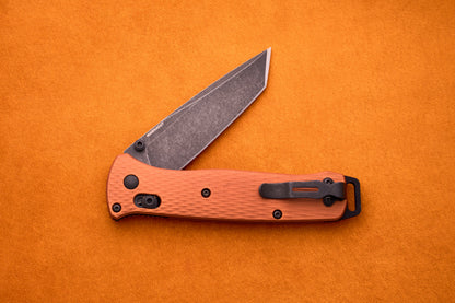 The Benchmade Bailout, featuring a textured orange handle and a partially open, gray CPM-Magnacut blade, is displayed on an orange background.